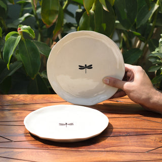 Hand holds off-white ceramic plate with black hand-painted dragonfly motif; second plate lies flat on wooden table with plants. Shop custom ceramics Bangalore.