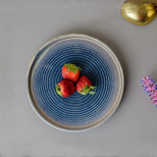 Small round blue glaze ceramic plate with circular pattern on interior, few strawberries in center on grey table. Buy ceramics for Indian homes.