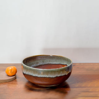 Large ceramic bowl glazed in earthy tones of brown and green inside and out, on a wooden table with an orange. Buy tableware online in India.