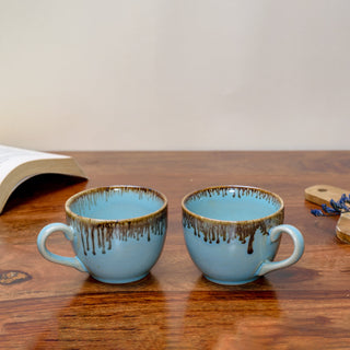 Set of small light blue ceramic cups with handle, brown drip glaze from rim, on wooden table, book, dried flowers in background. Buy traditional Indian ceramics.