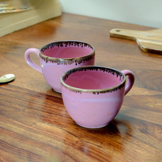 Close-up of 2 small ceramic pink cups with handles, drip brown glaze at rim, on wooden table, wooden slab, and book. Buy traditional Indian ceramics with Oh Yay Project