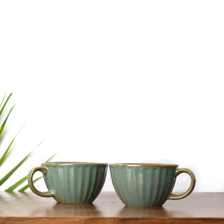 Pair of broad round green glaze ceramic cups with handles & brown glaze hints, on wooden table with green leaf in background. Shop handcrafted ceramics Bangalore.