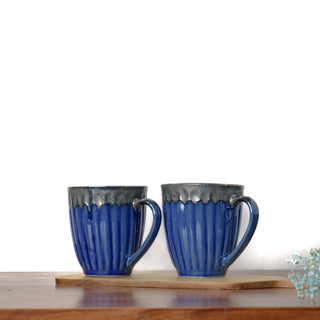 Pair of tall blue ribbed ceramic mugs with shiny glaze drip from rim and handle on wooden slab with blue dried flowers. Buy tableware online in Bangalore