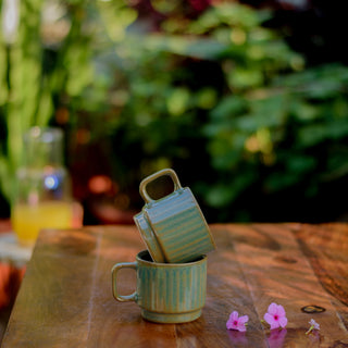 Pair of green glaze ceramic mugs: one on wooden table, other tilted on top with pink flowers, blurry greenery background. Buy kitchenware India.