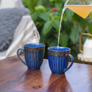 Pair of tall blue glaze mugs with tonal ribbed detailing, shiny drip glaze from rim, orange juice poured into one, on wooden table with blurry grey cushion and plants. Buy luxury ceramics Bangalore.