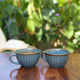 A pair of blue glaze broad round cups with handles, one with coffee, on wooden table with blurry book & plants in background. Buy tableware online India.