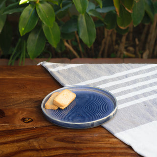 Small round blue glaze ceramic plate with circular interior design, 2 biscuits on half wooden table, half grey stripe throw, planT in background. Buy tableware online India.