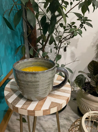 Large ceramic cup with handle in green earthy glaze filled with soup on round wooden table, plants in background. Buy tableware online India.