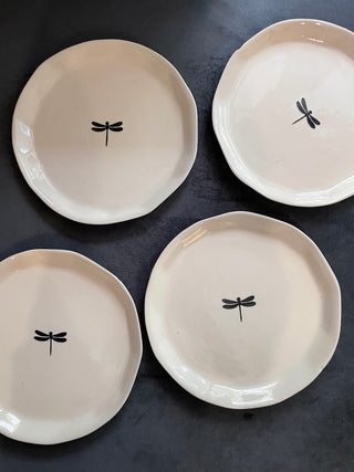 Top view of 4 uneven hem off-white ceramic plates with hand-painted black dragonfly motif on grey couch. Buy tableware online in Bangalore.