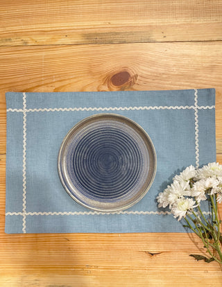 Rectangular blue table mat with silver scallop border creates check pattern near corners, with blue round plate and white flowers. Shop tableware online India