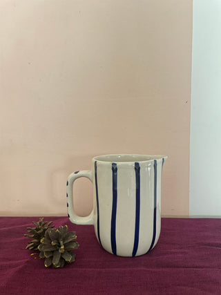 Small off-white ceramic jug with handle, hand-painted thin blue stripes kept on purple cloth with pine cones. Explore unique gifting ideas Bangalore.