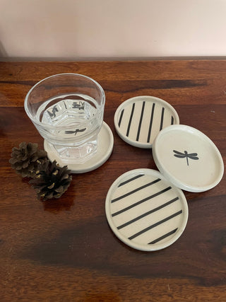 Set of 4 off-white coasters: 2 with black dragonfly motifs, 2 with black stripes, one with a glass of water and pine cones on a wooden table. Tips on styling your home with ceramics.