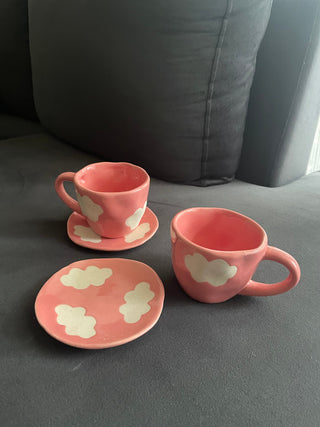 Set of 2 pink cup saucers with white cloud motif in stoneware finish: one intact, one cup outside saucer, on grey sofa. Shop luxury ceramics Bangalore.