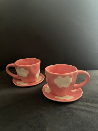 Set of 2 pink cup saucer sets with cute cloud motifs in stoneware finish, placed on grey sofa. Explore tableware online India.