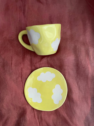 Top shot of yellow cup & saucer with cloud motifs in stoneware finish placed flat on a maroon cloth. Explore custom ceramics Bangalore.