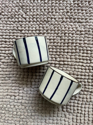 2 small white ceramic tea cups with thin blue hand-painted stripes, brown rim, on grey bubble fabric. Buy tableware for special occasions India.