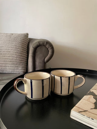 Pair of small off-white ceramic tea cups with thin blue hand-painted stripes, brown rim, and handle on round table next to a book and grey couch. Explore kitchenware India