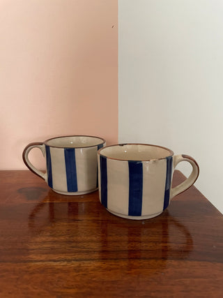 Pair of small ceramic tea/coffee cups: off-white with broad blue stripes, goldish brown rim and handle, on wooden table. Shop unique gifting ideas Bangalore.