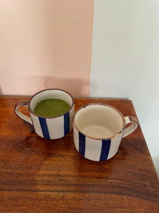 Top shot of small ceramic tea cups: off-white with broad blue stripes, one filled with matcha, the other empty. Explore traditional Indian ceramics.