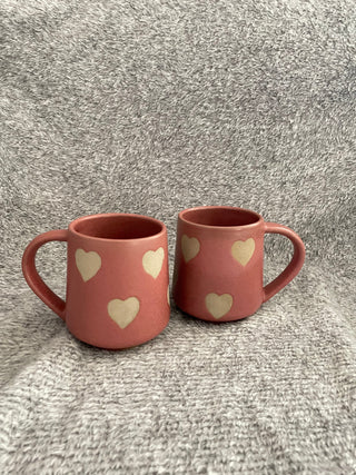 A pair of tall pink ceramic mugs with handles, adorned with heart motifs in stone ware finish, on a soft grey fabric. Buy online ceramics store India