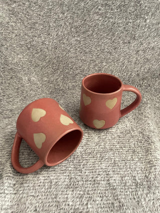 Set of pink medium ceramic mugs with handles adorned with beige hearts, one flat and the other standing on grey fabric. Buy handcrafted ceramics in India