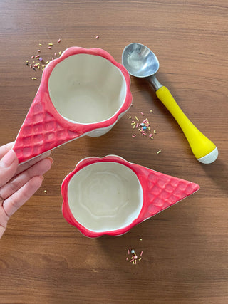 Pair of unique red glazed ceramic ice cream bowls, one held up by hand, sprinkles on table, yellow scooper.