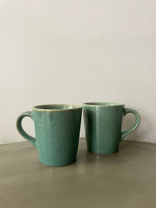 Pair of beautiful teal glaze medium tall ceramic mugs with handles, placed on cement table. Shop tableware online in India.