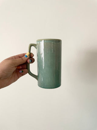 Hand with vibrant painted nails holding tall ceramic mug, long handle for easy use, finished in teal glaze. Shop handcrafted ceramics in India.