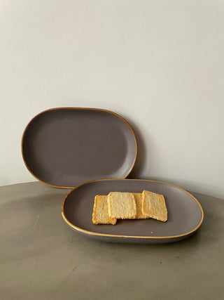 A set of 2 small oval grey stoneware finish tapa plates with brown rim, one against a wall, other flat on cement table with biscuits. Buy tableware online in India.