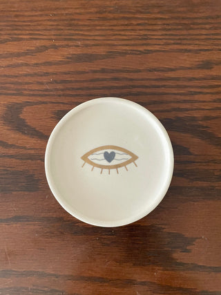 Small round off-white coaster with gold evil eye and blue heart as eyeball, placed on wooden table. Explore Oh Yay Project's online ceramics store India.