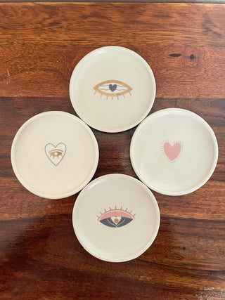 Set of 4 small round off-white coasters: 2 evil eye, 2 hearts in blue, pink, gold, placed flat on wooden table. Explore handcrafted ceramics India.