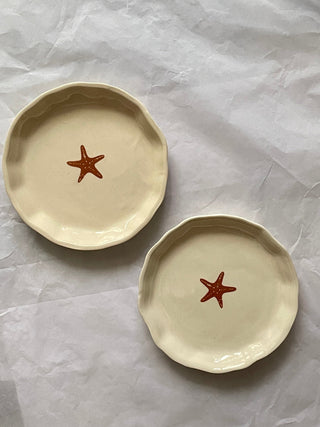 Set of 2 small uneven hem off-white ceramic plates with orange starfish motif on white sheet. Buy handcrafted ceramics in India.