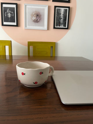 A big round white ceramic mug with uneven rim, hand-painted red cherry and white swirl design, short handle on dining table with laptop. Buy tableware online Bangalore.