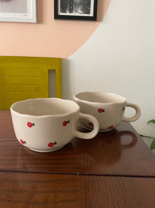 Big broad uneven hem white ceramic mugs with hand-painted cherry swirl designs, short long white handle, on dining table. Buy artistic ceramics in Bangalore.