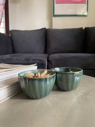 Close-up of 2 green glazed ceramic bowls on a cement table with 2 books; one bowl has snacks, the other is empty. Shop at Oh Yay Project's online ceramics store in India.