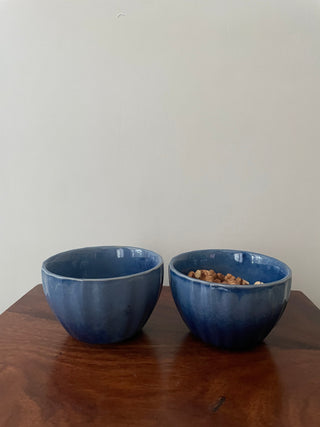 A set of 2 small blue ceramic bowls on a wooden table. One bowl is filled with walnuts, the other is empty. Shop perfect ceramics for Indian homes.