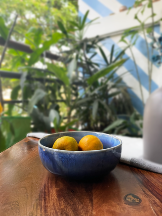 Medium blue glaze ceramic bowl with oranges placedon wooden table with blury plants background. Buy tableware online in India