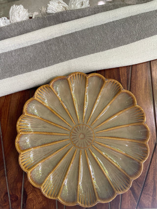 Large flower-shaped beige ceramic platter with mustard glaze line design and a mini flower, on a wooden table with a grey throw. Shop unique gifting ideas Bangalore.